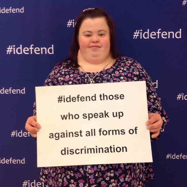 Woman holds poster #defend those who speak up against all forms of discrimination.