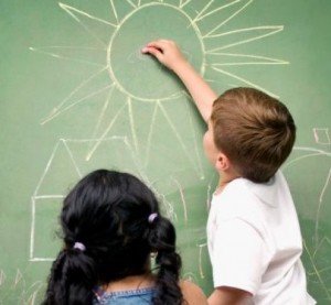 Menino desenha casa e sol na lousa e menina observa, ambos de costas para a camera Menino desenha casa e sol na lousa e menina observa, ambos de costas para a camera