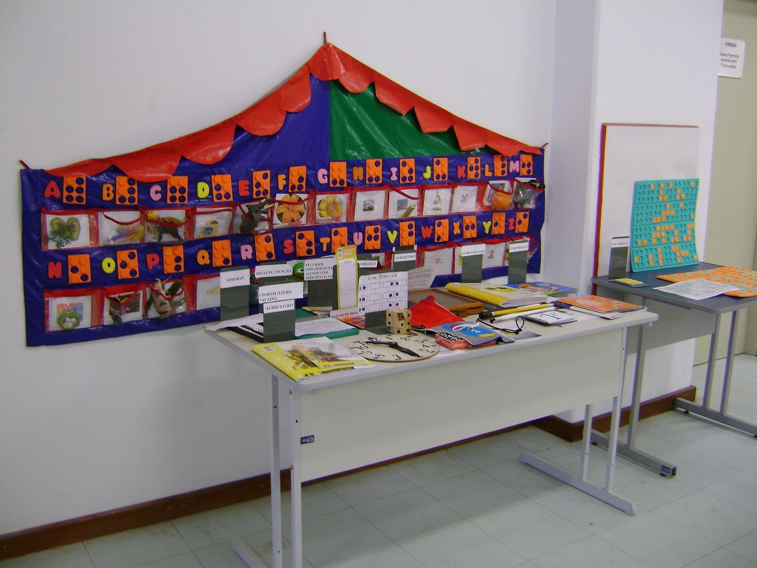 Material tatil para ensino de Braille disposto numa tenda colorida presa na parede. Mesa com recursos para ensino de pessoas com deficiência visual.