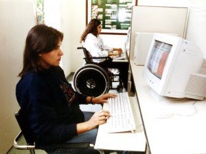 Duas mulheres sentadas ao computador, trabalhando, uma delas, em segundo plano, sentada em cadeira de rodas.