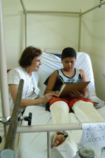 educacao hospitalar Menino com duas pernas engessadas lê livro ao lado de professora