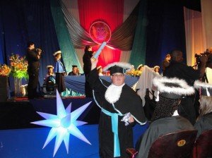 Foto: Gustavo Bicca, em primeiro plano, de pé, segurando o diploma com o braço direito erguido, como se portasse uma espada; a mão esquerda está na cintura. Sua pose é a de um vitorioso. Como os demais, ele veste uma toga preta, longa, com uma faixa azul na cintura; na cabeça, o chapéu, que tem o nome de "capelo" e arminho nas bordas. Ao fundo, a mesa com as autoridades.