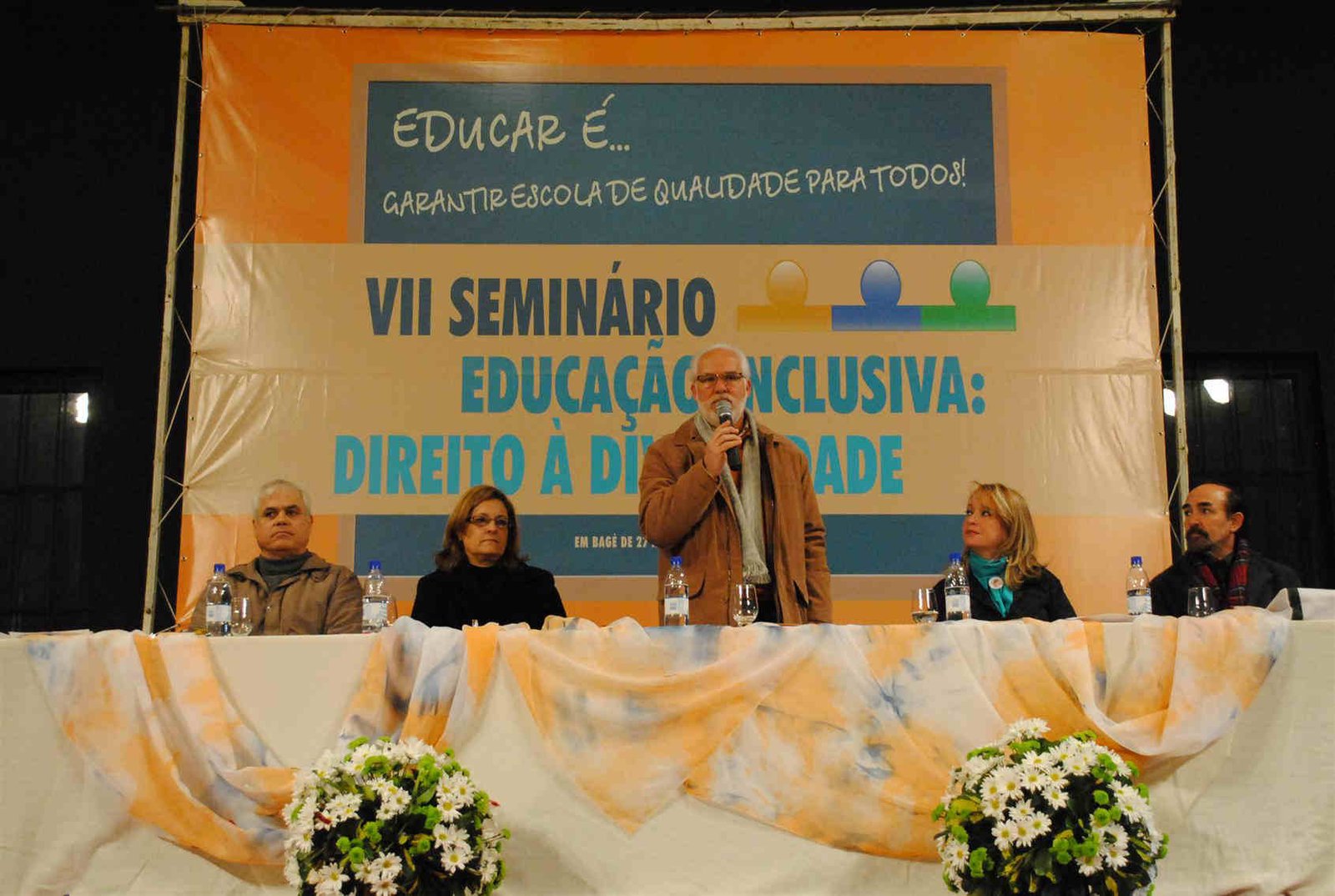 27_06 Quatro palestrantes sentados à mesa com dois arranjos de flores à frente e o banner do encontro ao fundo e no centro um palestrante de pé falando ao microfone.