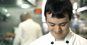 Rapaz com sindrome de Down vestindo uniforme de cozinehiro olha para baixo e sorri. Descricao do video com legendas em ingles - em uma cozinha de restaurante, Cena de frigideira saindo labaredas no fogao, imagem em close de vegetais e carne sendo picados com faca em uma tabua. Camera mostra que quem pica e um rapaz com sindrome de Down e roupa de cozinheiro. Chef principal, calvo, em frances, diz que o restaurante esta cheio e que ele precisa que todos os funcionarios deem 200% de si mesmos. Virando-se para o rapaz com sindrome de Down diz : " vamos la, as pessoas estao esperando, o que deu em voce hoje?" . A camera abre revelando um outro ajudante de cozinha, sem deficiencia, e o Chef diz: " E voce? Ve se acorda!" , e sai. O ajudante de cozinha, balanca a cabeca, chateado com a bronca. O rapaz com sindrome de Down, olha para baixo e sorri.  Serge o letreiro: OBRIGADO. Por nos ratar como gente normal..