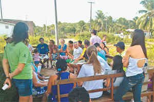 Grupo de pessoas, algumas sentadas, outras de pé participa de debate ao ar livre.