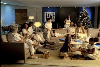 Em uma sala, com arvore de natal ao fundo, varias pessoas vestindo branco sentadas num sofa, meninas brincando sentadas no chao.