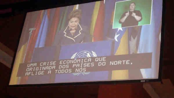 Imagem da projeção em telão da Presidente Dilma Rousseff, com legenda e janela de libras aparente