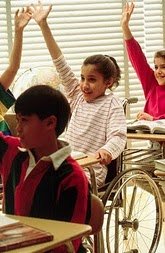 Em sala de aula, sentados as carteiras alunos levantam a mão - na primeira carteira um menino com síndrome de Down, na segunda uma menina cadeirante, na terceira uma menina sem dificiência.