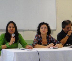 Sentados à mesa do evento, três ativistas, 1 homem e duas muljheres, uma delas falando ao microfone.