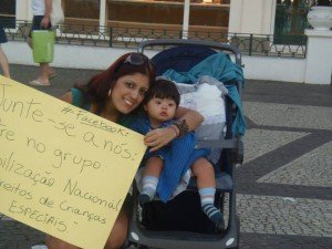 Manifestante abaixada junto ao carrinho da filha, segura cartaz dizendo - Junte-se a nós no Facebook - Mobilização Nacional pelos Direitos das Crianças Especiais.