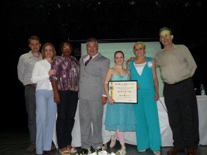 Aline, com roupa de bailarina, segura o título, junto a familiares e autoridades.