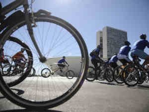 Várias ciclistas pedalando, com uma roda de bicicleta em primeiro plano, no fundo o ceu azul e os prédios do Congresso Nacional.