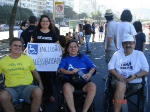 Três manifestantes em cadeira de rodas e uma com a camiseta Ser Diferente é Normal, mostrando um cartaz que diz Inclusão e Acessibilidade.