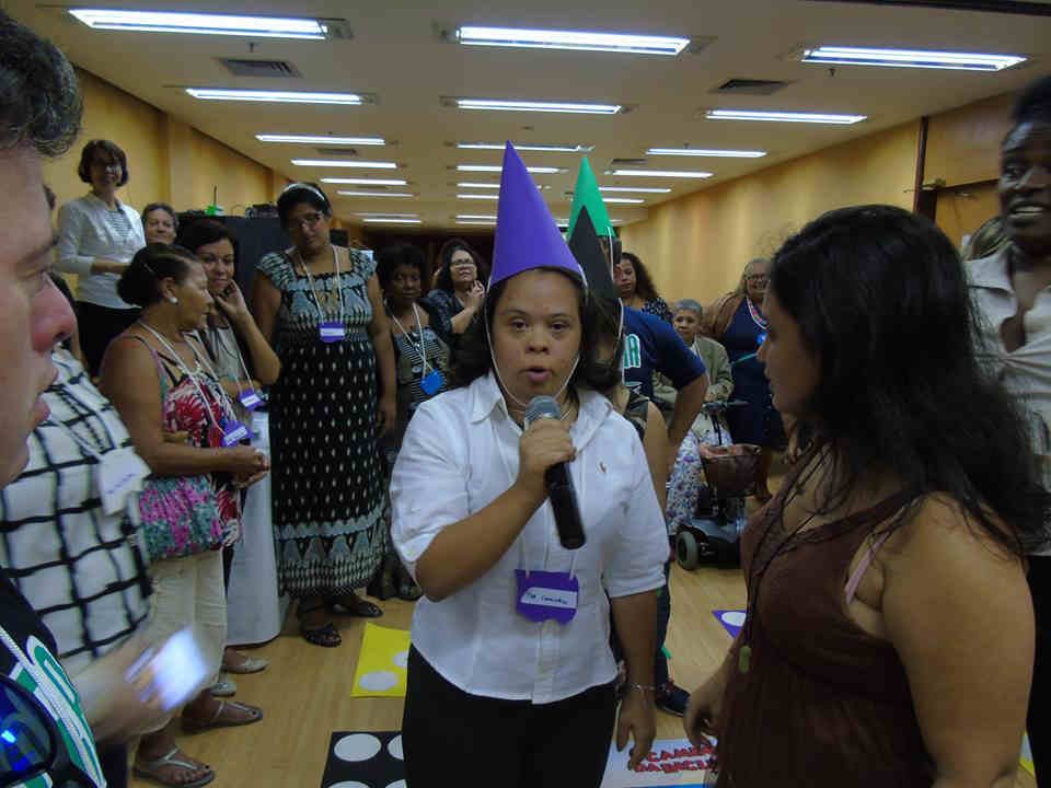 jovem participante da mostra fala ao microfone em sala, em meio a outros participantes. ela tem chapeu em cone roxo.