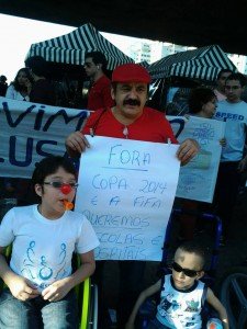 Na manifestação, homem de camisa e bone vermelho, de bigode, carrega cartaz "Fora Copa 2014 e a Fifa. Queremos Escolas e Hospitas". Está entre dois meninos de cadeira de rodas, um deles com nariz de palhaço.