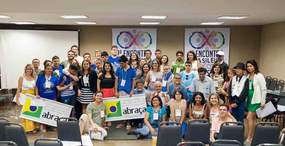 varias pessoas em uma sala, algumas de pe, outras sentadas no chao, com banners da abraça e do I encontro de pessoas autistas.