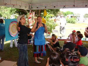 Contadora de histórias de avental azul, ao lado de intérprete de Libras, de camiseta preta e boné bege, contam história para grupo de crianças sentadas no chão em tenda ao ar livre.