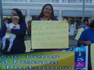 Mulher segura cartaz dizendo - Necessitamos maior agilidade no agendamento de consulas, exames e tratamentos., ao lado dela, moça com bebê com síndrome de Down no colo.