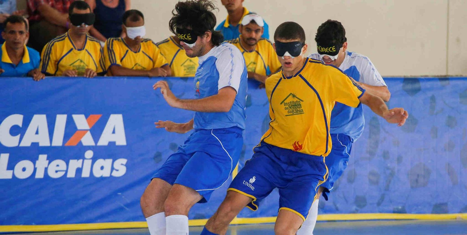 dois jogadores de futebol em acao. eles usam vendas. um tem uniforme azil e branco e o outro amarelo e azul. ao fundo, jogadores que estão no banco acompanham, alguns com e outros sem venda.