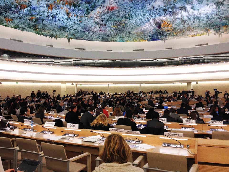 sala do conselho dos direitos humanos da ONU, com pessoas sentadas em circulo e o teto colorido.