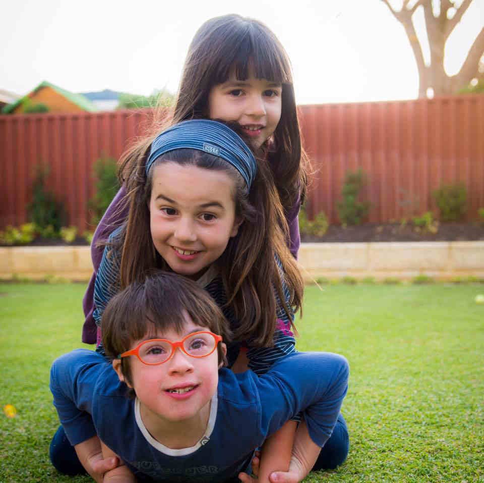 tres criancas, uma em cima da outra num gramado. um menino de oculos vermelhos, com sindrome de down por baixo, uma menina por cima e outra menina por cima.