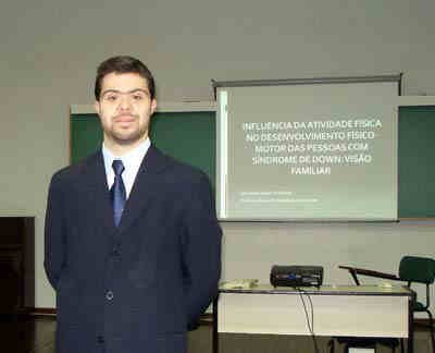 João Victor, que tem síndrome de Down, diante de uma tela com a apresentação de seu trabalho final na universidade " Influência da atividade física no movimento motor das pessoas com síndrome de Down. Visão familiar".