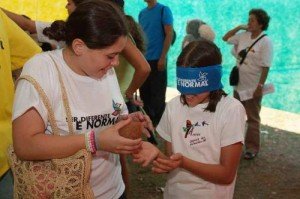 Mulher com camiseta Ser Diferente é Normal passa coco seco para a mão de menina com uma venda nos olhos.