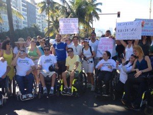 Diversas pessoas com e sem deficiência, algumas em cadeiras de rodas, mostram cartazes.