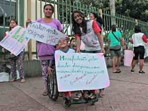 Criança em cadeira de rodas segura cartaz ao lado de duas mulheres, uma delas também empunhando cartaz.