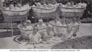 Foto em preto e branco mostrando 5 berços antigos com cerca de 4 bebês em cada, e três sentados no chão.