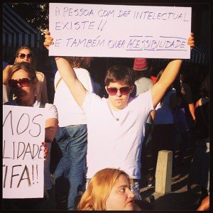 Jovem de óculos escuros segura cartaz - a pessoa com deficiência intelectual existe e também quer acessibilidade.