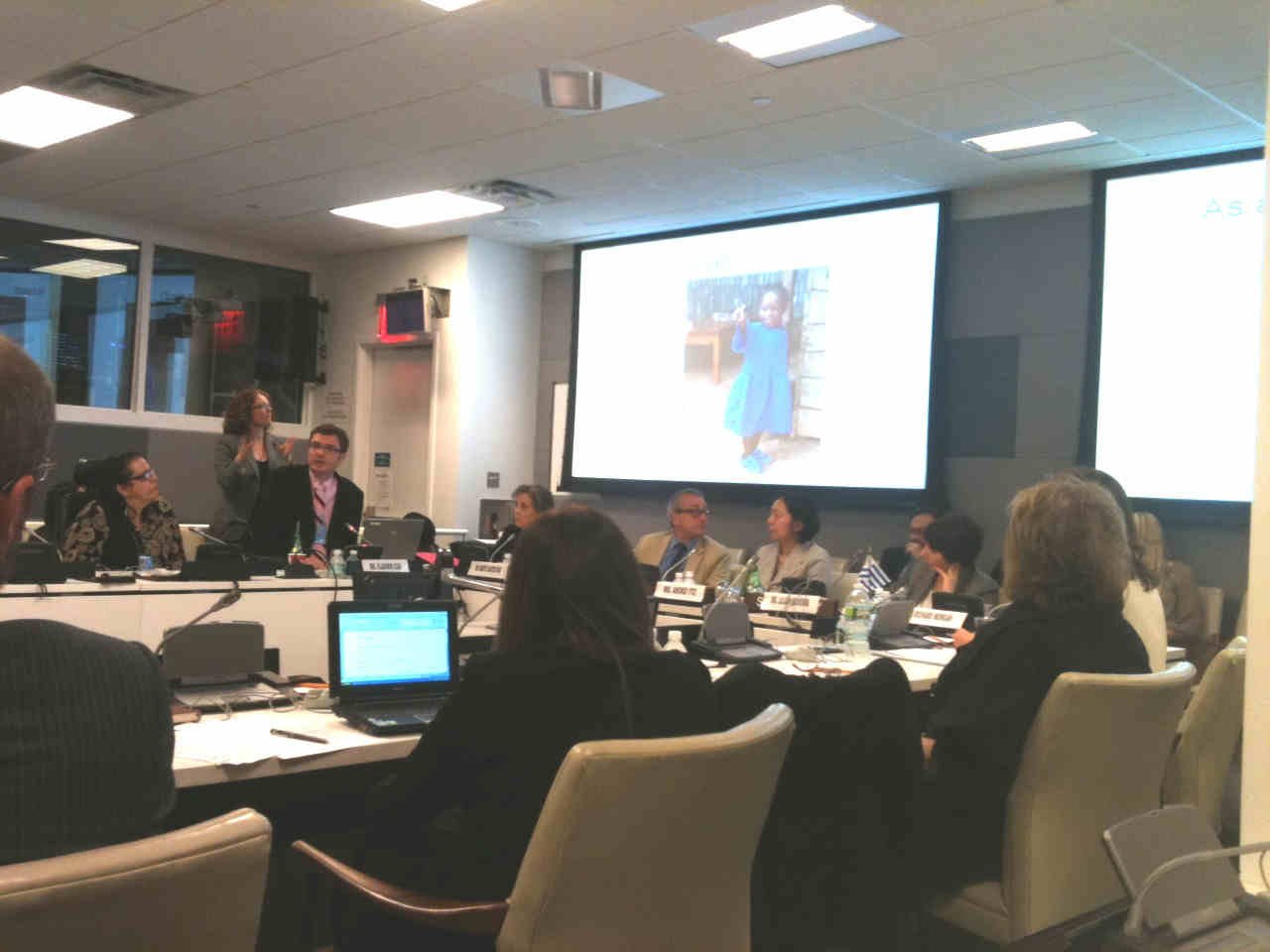 Sala de reunião com várias pessoas sentadas à volta da mesa e apresentação sendo projetada em tela, mostrando menina pequena com vestido azul.