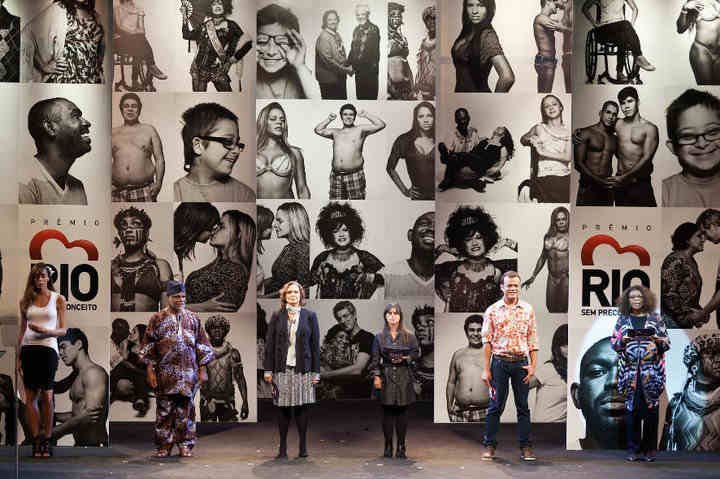 6 pessoas no palco diante de painel de fotos em preto e branco de pessoas alvo de preconceito. 6 pessoas no palco diante de painel de fotos em preto e branco de pessoas alvo de preconceito.