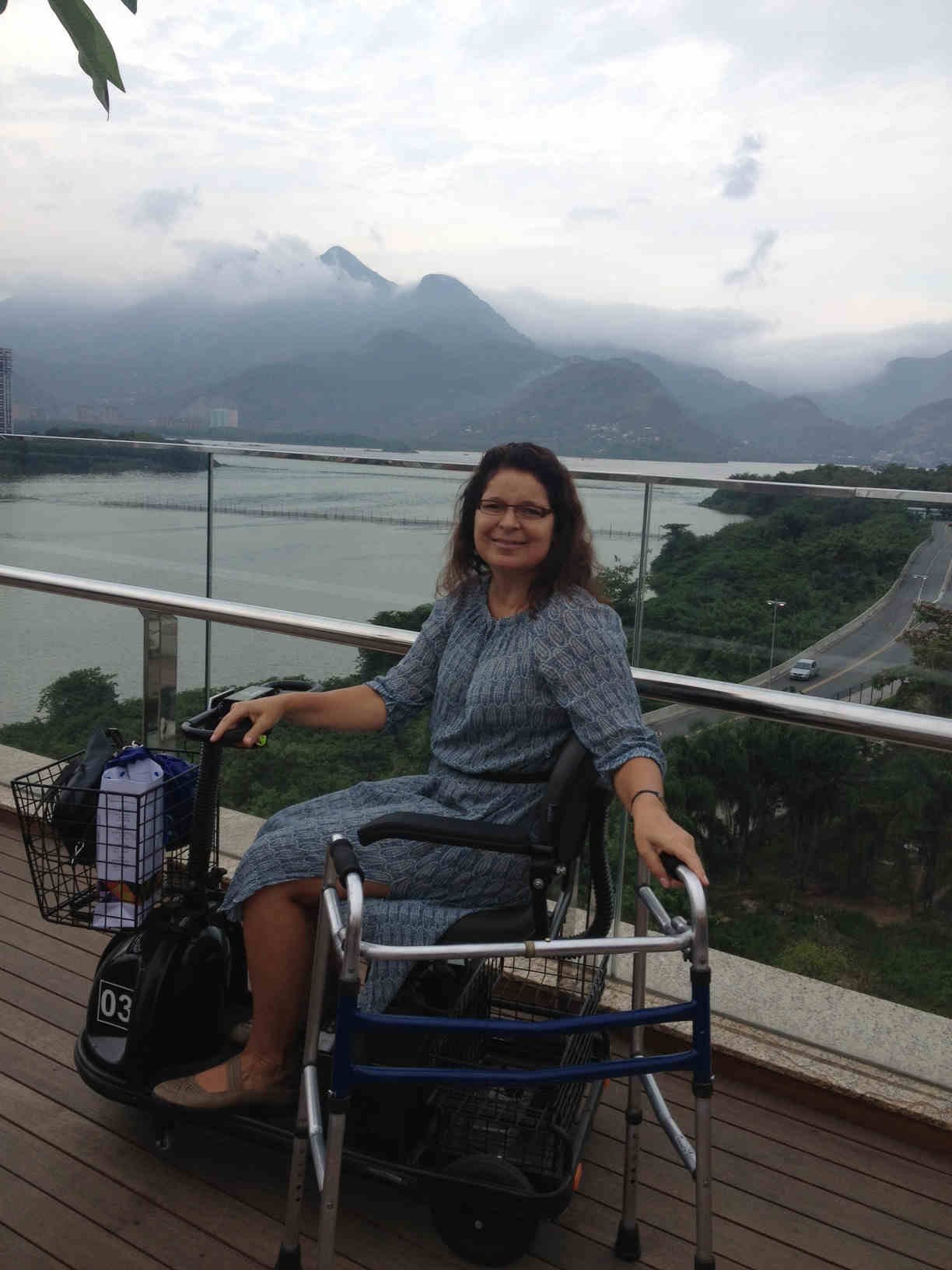 Patricia na cadeira motorizada com o andador ao lado no terraço do Village Mall, com a paisagem da Lagoa da Tijuca e montanhas ao fundo.