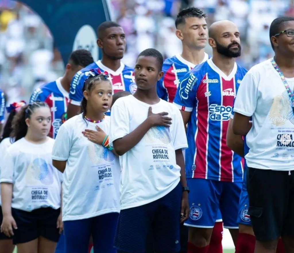 duas moças e um rapaz com a camiseta chega de rotulos, dois deles com a mao no peito, ao lado de jogadores em estadio de futebol na hora do hino nacional.