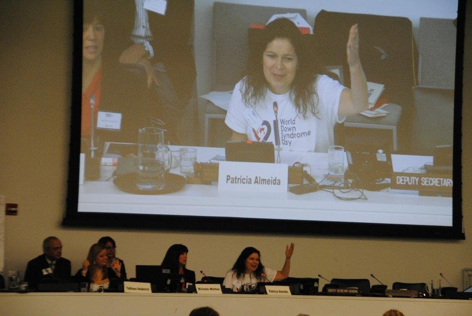 mulher sentada à mesa de auditório com camiseta do dia internacional da síndrome de Down, placa esccrito Patricia Almeida, com a mão levantada e sua imagem na tela acima