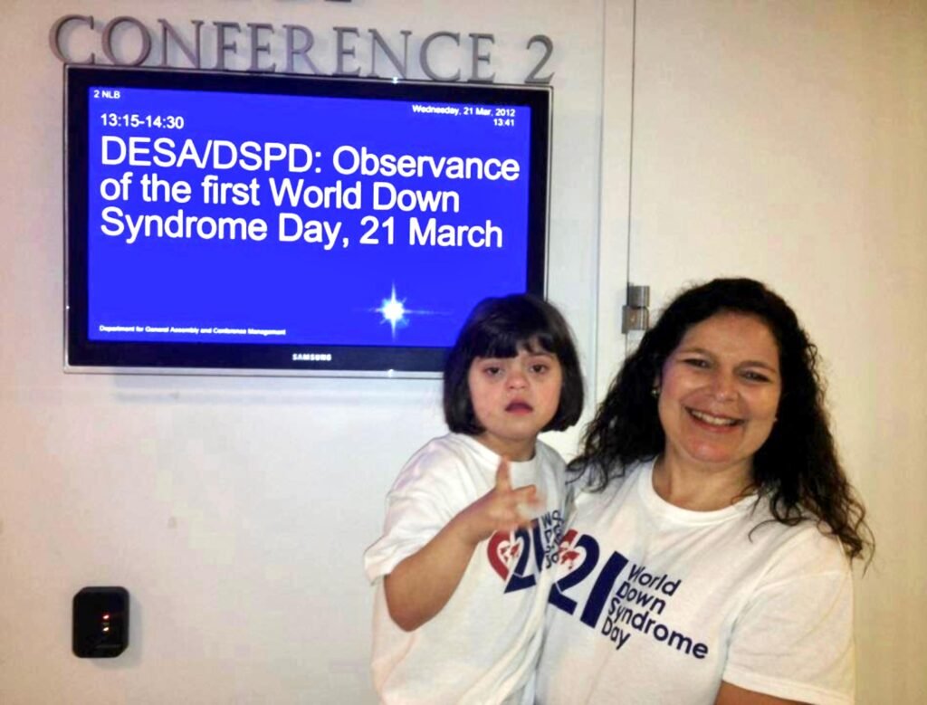 mulher com menina no colo em frente a tela indicando evento sobre o dia internacional da sindrome de down, salão de conferências 2.
