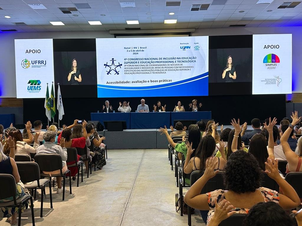 sala de conferências, telão, público batendo palmas em siléncio, com as mãos para cima.