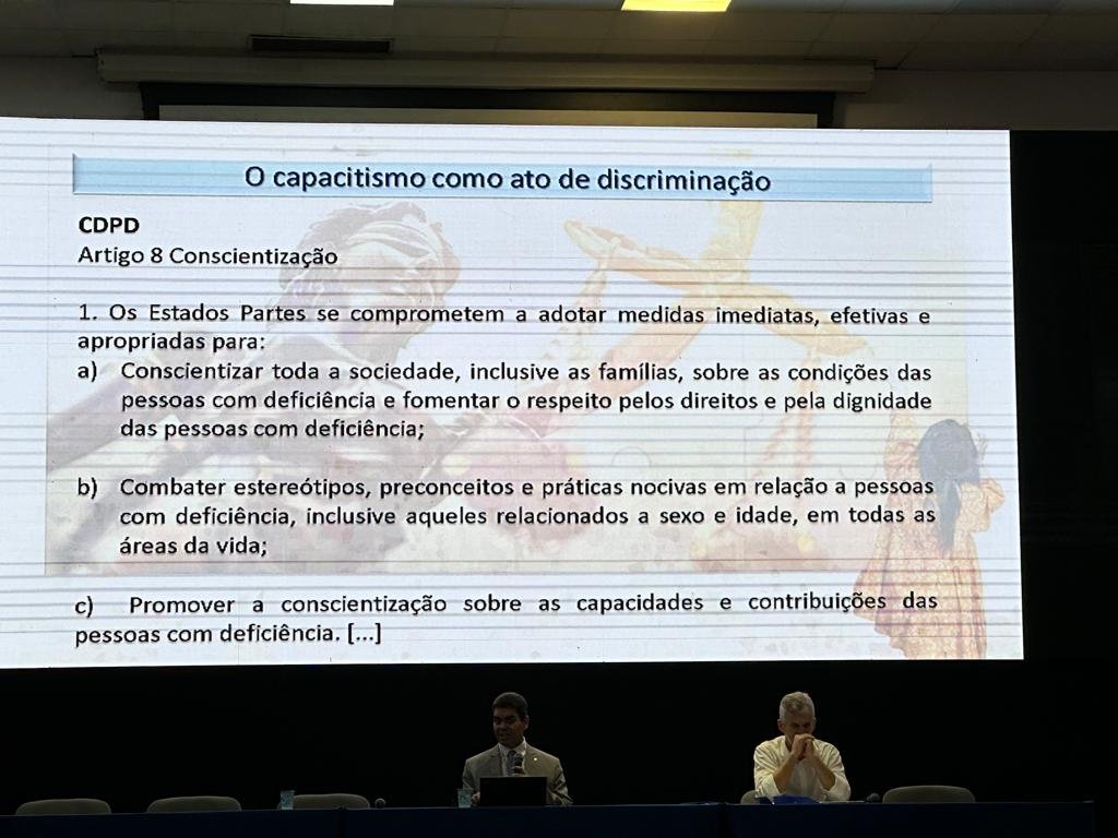 tela com slide com título capacitismo como ato de discriminação, mesa com dois homens sentados.