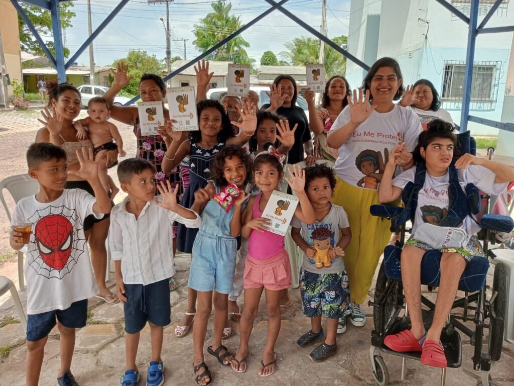 grupo de várias crianças com a cartilha eu me protejo e a mão espalmada à frente, entre eles menina em cadeira de rodas e mulher com camiseta eu me protejo.