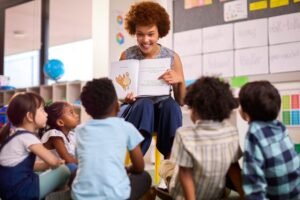 Uma professora com cabelo afro e um grande sorriso lê um livro ilustrado para um grupo de crianças sentadas em círculo ao seu redor. O ambiente é uma sala de aula colorida e bem iluminada, com cartazes e materiais educativos nas paredes.