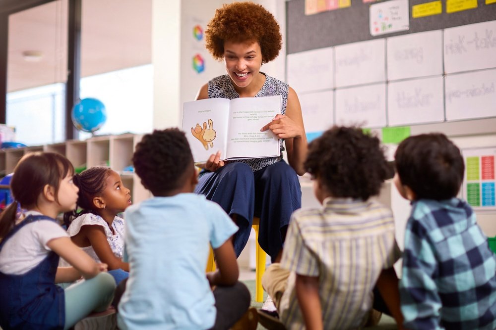 Uma professora com cabelo afro e um grande sorriso lê um livro ilustrado para um grupo de crianças sentadas em círculo ao seu redor. O ambiente é uma sala de aula colorida e bem iluminada, com cartazes e materiais educativos nas paredes.