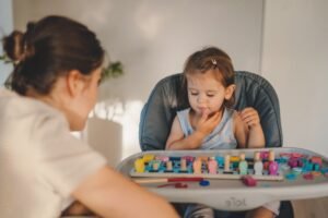 A imagem mostra uma criança pequena, uma menina, sentada em uma cadeira alta para bebês. Ela está brincando com um brinquedo educativo colorido que envolve peças de madeira de diferentes formas e cores. A criança está concentrada, observando o brinquedo. À esquerda, uma mulher de cabelo preso, parcialmente fora de foco, parece interagir com a criança, possivelmente oferecendo apoio ou instruções. O ambiente é iluminado e acolhedor, sugerindo um momento de aprendizado ou brincadeira.