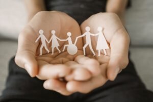 A imagem mostra duas mãos abertas segurando recortes de papel em formato de figuras humanas. As figuras formam uma cadeia de pessoas de mãos dadas, incluindo uma pessoa em uma cadeira de rodas no centro. A cena simboliza inclusão, apoio e união. As figuras humanas são simples, estilizadas, e a composição geral transmite uma mensagem de solidariedade e acolhimento para todas as pessoas.