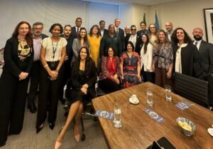 A imagem mostra um grupo de aproximadamente 20 pessoas reunidas em uma sala de conferências da ONU. Há uma mistura de homens e mulheres de diferentes idades e etnias, todos sorrindo para a câmera. Três mulheres estão sentadas em cadeiras na frente do grupo, uma delas é Anna Paula Feminella, Secretaria dos Direitos da Pessoa com Deficiência do Ministério dos Direitos Humanos e da Cidadania, diversas pessoas estão de pé atrás delas. A sala tem uma mesa grande com alguns copos de água e xícaras de café. Ao fundo, há duas bandeiras, uma do Brasil e outra da Organização das Nações Unidas.