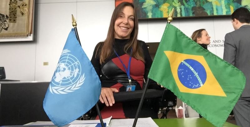 foto de mara gabrilli, em cadeira de rodas, a frente de duas bandeiras, do brasil e da onu.