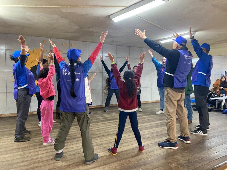 A imagem mostra um grupo de adultos e crianças participando de uma atividade em grupo dentro de uma sala. As pessoas estão formando um círculo com os braços levantados, como se estivessem se alongando ou fazendo algum tipo de exercício. Os adultos estão usando coletes roxos com um logotipo nas costas e bonés azuis, sugerindo que são instrutores ou voluntários de alguma organização. As crianças estão vestidas com roupas casuais. A sala tem piso de madeira e paredes de azulejo, e parece ser um ambiente fechado e iluminado por lâmpadas fluorescentes no teto.