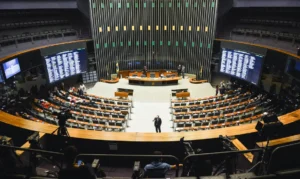 A imagem mostra uma grande câmara legislativa com assentos dispostos em semicírculo voltados para um pódio central. Há grandes telas eletrônicas em ambos os lados da sala, exibindo informações ou resultados de votação. Algumas pessoas estão presentes, algumas sentadas nos assentos e outras se movimentando pelo local.