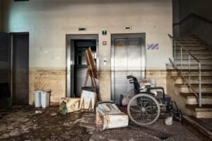 A imagem mostra uma cena de abandono em um edifício, possivelmente um hospital ou uma instituição de saúde. No centro da imagem, há dois elevadores; o da esquerda está aberto e bloqueado por um cavalete de madeira. Em frente aos elevadores, há diversos objetos danificados e cobertos de sujeira, incluindo uma cadeira de rodas e algumas caixas de armazenamento. À direita, há uma escada que parece ter sido afetada por uma enchente ou inundação, evidenciada pelo nível de sujeira nas paredes e no chão. O ambiente está sujo, desorganizado e parece ter sido evacuado ou abandonado há algum tempo.