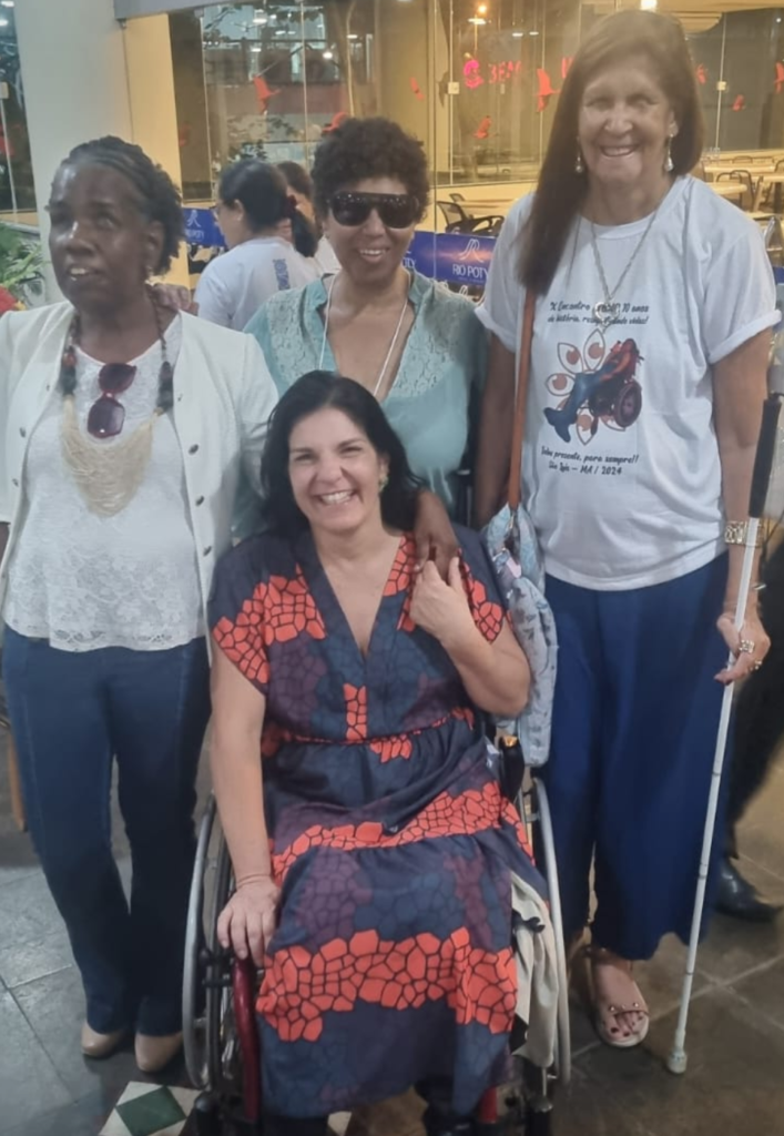 3 mulheres, uma com bengala branca, em pé e uma mulher em cadeira de rodas, todas sorrindo.